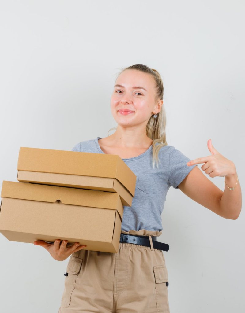 Young lady pointing at cardboard boxes in t-shirt, pants and looking merry. front view.